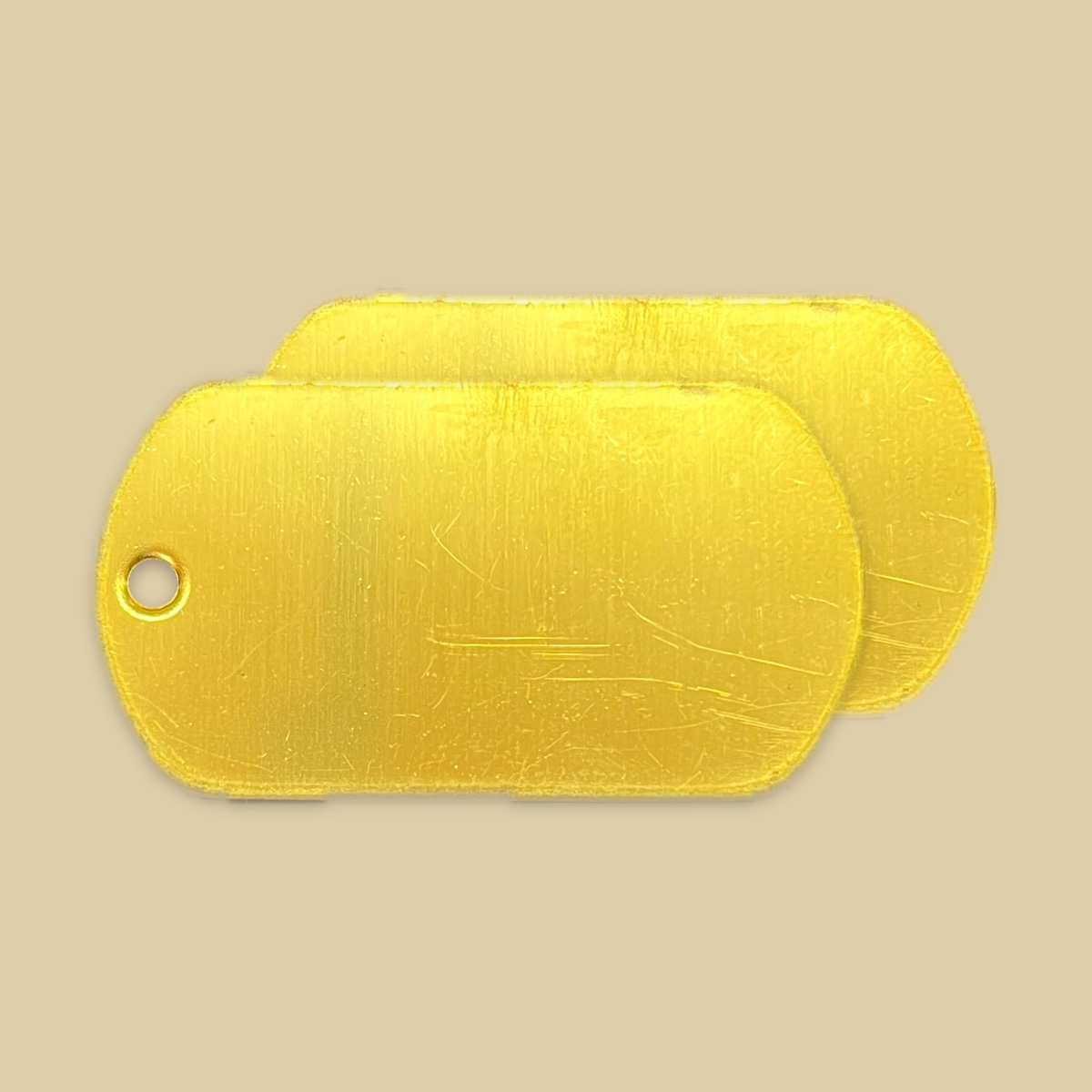 Two blank limited edition brass tags on a plain khaki backdrop, one lying over the other and offset so both are visible. The faces are a warm polished gold with a vertical brushed grain and a few light scratches, and the front tag shows its bead chain hole at the left end.