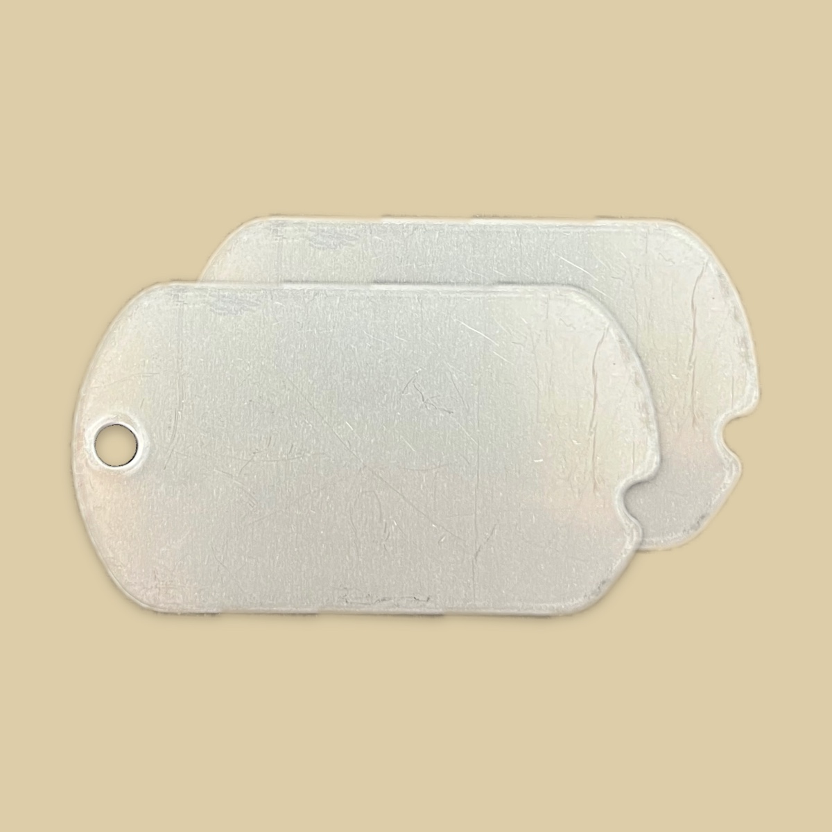 Two blank notched WWII replica tags on a plain khaki backdrop, one lying over the other and offset so both are visible. The brushed steel faces carry fine scratches, a notch is cut into the middle of the short right edge of each tag, and the front tag shows its bead chain hole at the left end.