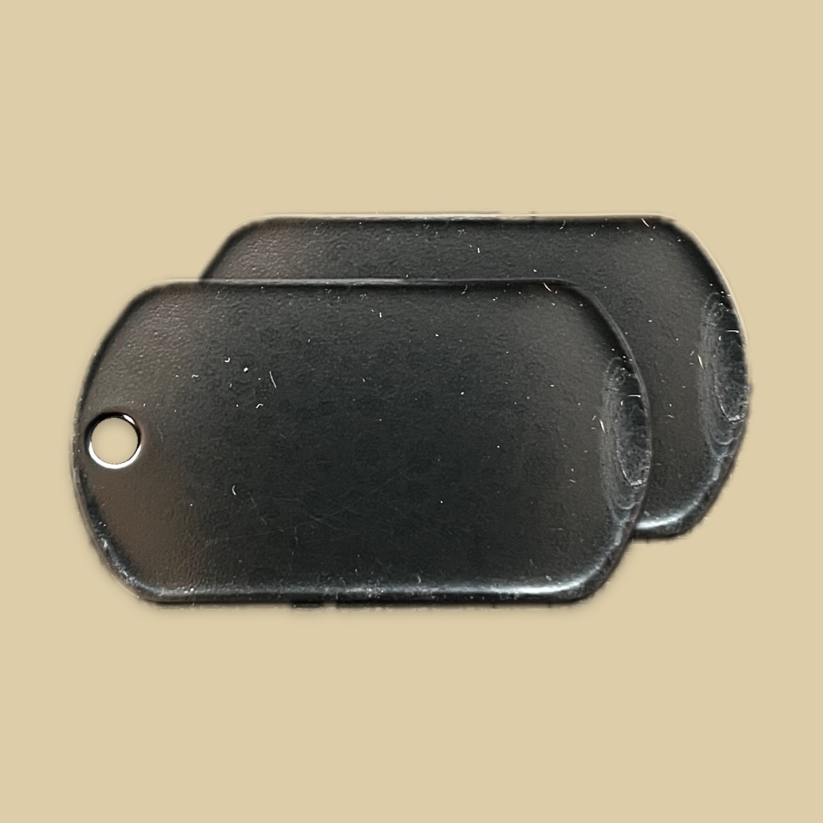 Two blank tags in the polished stainless and black finish on a plain khaki backdrop, one lying over the other and offset so both are visible. The faces are a deep glossy black that catches a soft highlight along the top edge, and the front tag shows a bare metal bead chain hole at the left end.