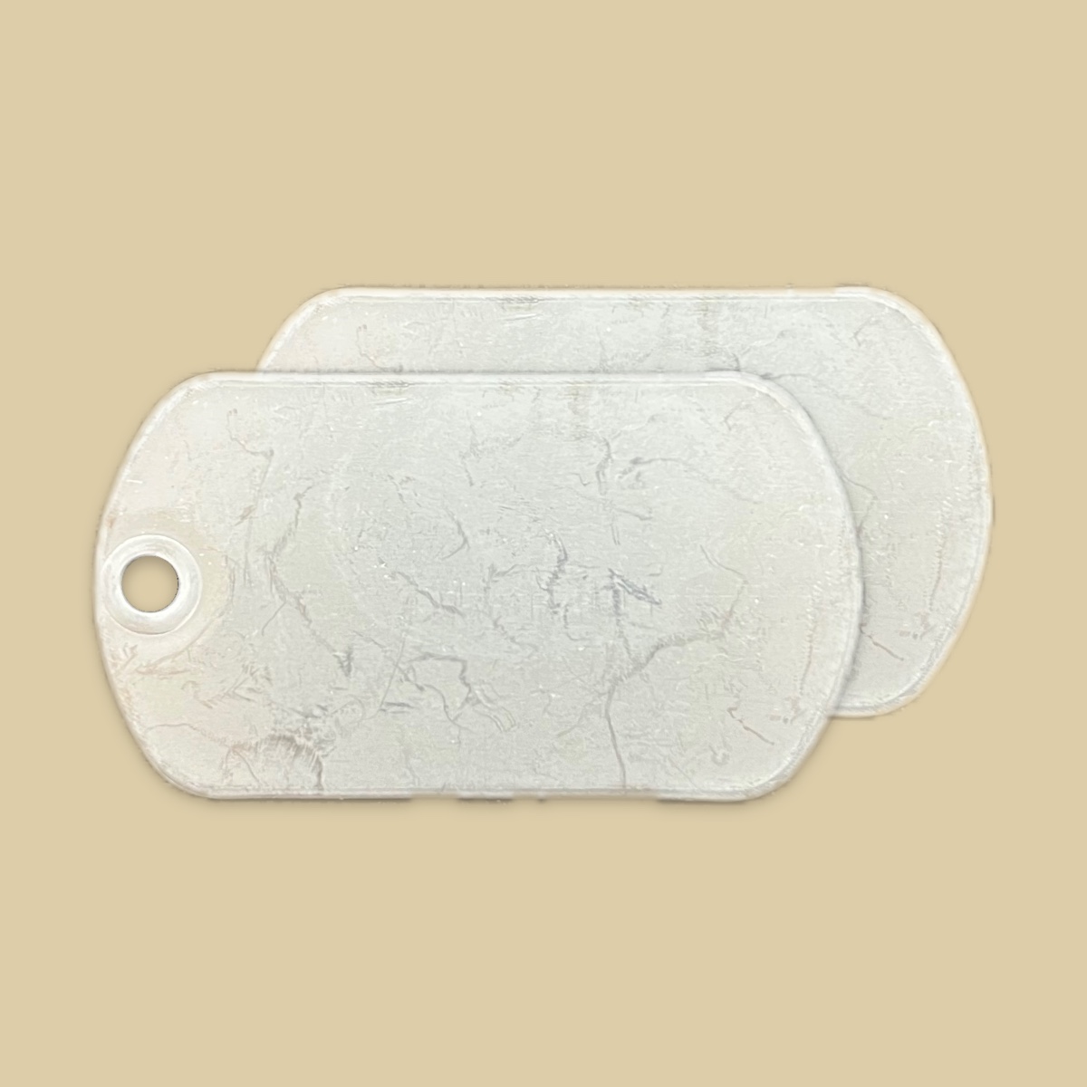 Two blank standard stainless steel tags on a plain khaki backdrop, one lying over the other and offset so both are visible. The faces are a dull unpolished grey with a mottled patina and fine scratches, and the front tag shows its bead chain hole at the left end.